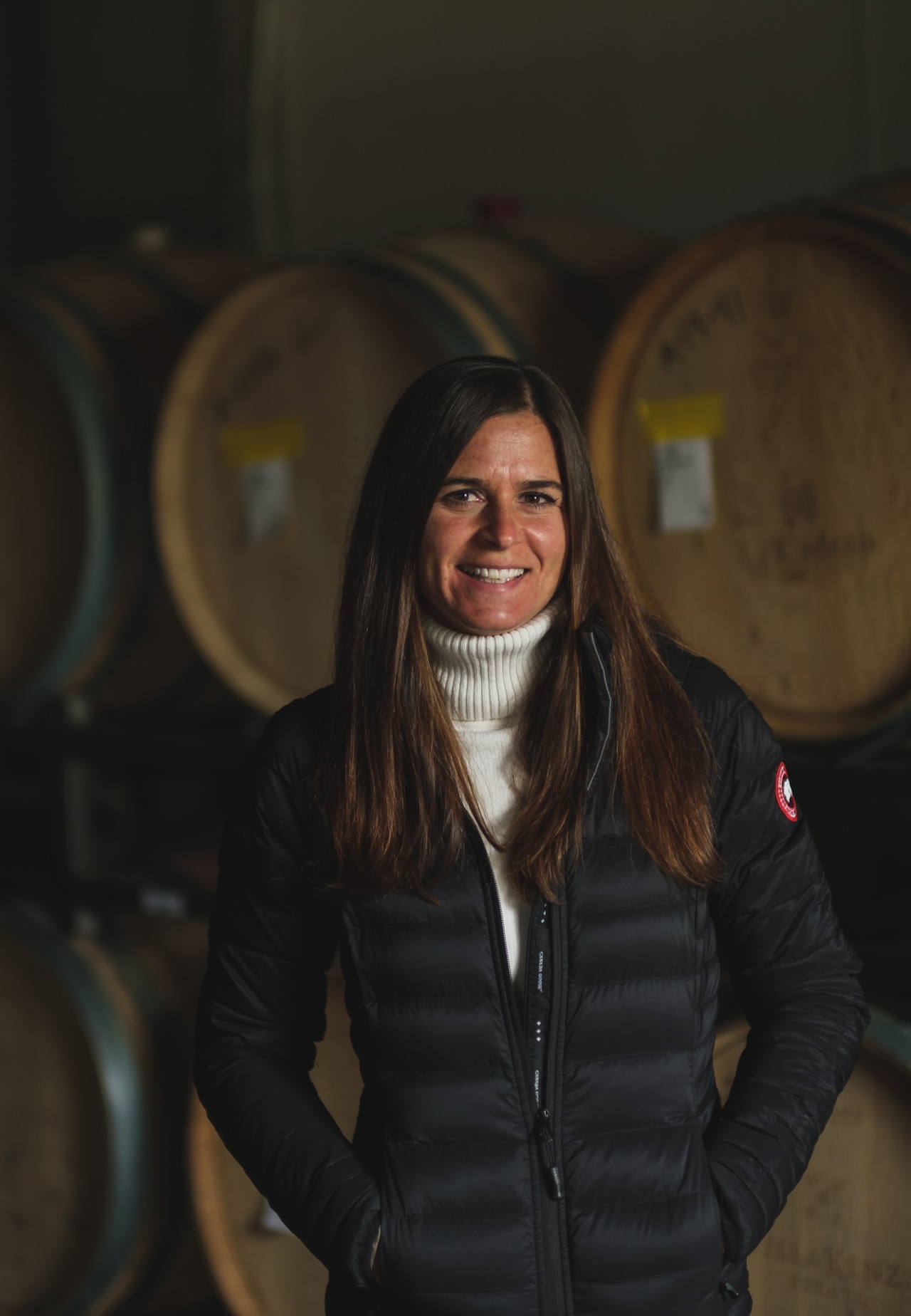 A woman wearing a puffer jacket stand in front of oak wine barrels.