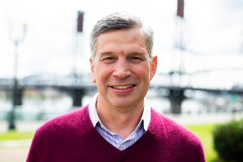 A man with grey hair wearing a burgundy pullover stands facing the camera and smiling