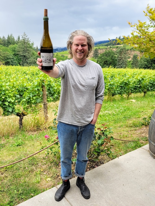 A man holding a bottle of wine stands in front of a vineyard.