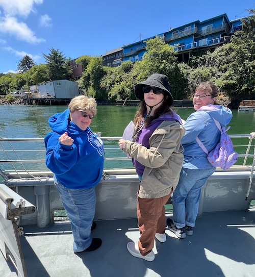 Three people are shown posing for a photo next to the water as they lean on a railing