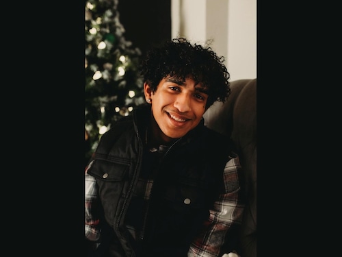 An undated family photo of Asher Ethan HK Mendes. He's wearing a black jacket with a gray and red flannel, smiling on a couch next to what appears to be a Christmas tree.