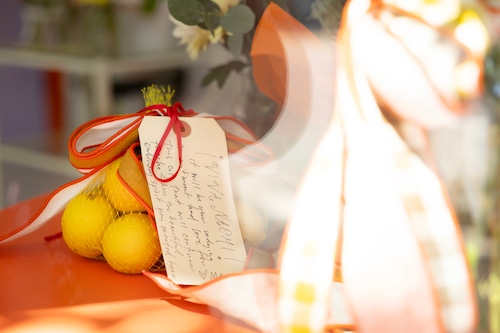 A bag of lemons sit with a note on an orange metal table next to a bouquet of flowers.