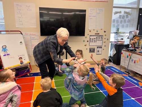 A woman in a blazer high-fives a group of young children seated on a rug in a classroom.