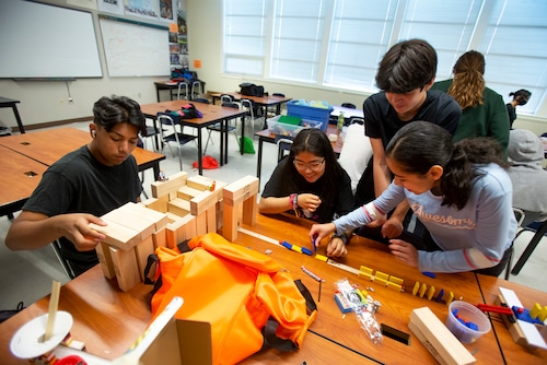 Four teenagers sit at or stand next to a desk, where colorful blocks are arranged on the surface in front of them