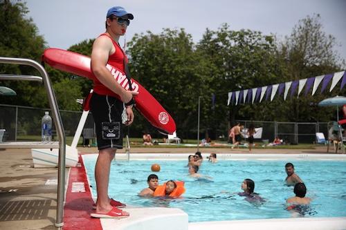 A life guard holding a red life preserver stands at the edge of an outdoor public swimming pool, overlooking a half-dozen swimmers.